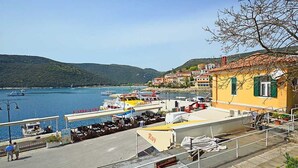 Una marina costiera con imbarcazioni ormeggiate, un edificio giallo con persiane verdi e uno sfondo collinare.