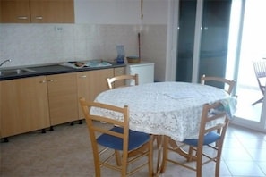 Eine Küche mit Holzausstattung, einem Spülbecken und einem kleinen Kühlschrank. Ein runder Tisch mit Stühlen und ein Fenster sorgen für natürliches Licht.