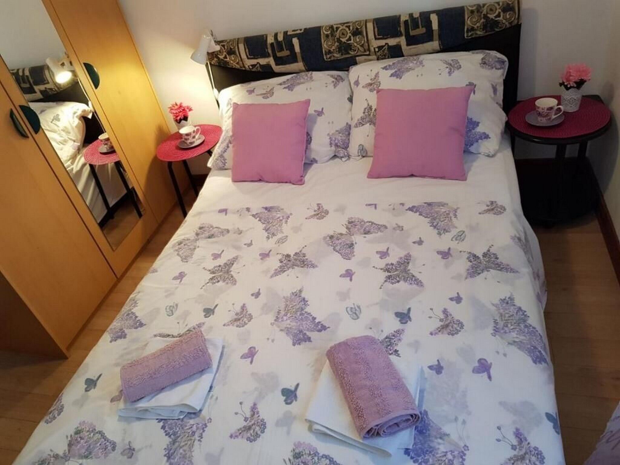 A neatly made bed with floral bedding, two pink pillows, and two folded towels. There are two bedside tables with lamps and a small vase of flowers on each.