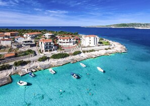 Vista aérea de un pueblo costero con edificios a lo largo de la costa, barcos en el agua y un cielo azul despejado.