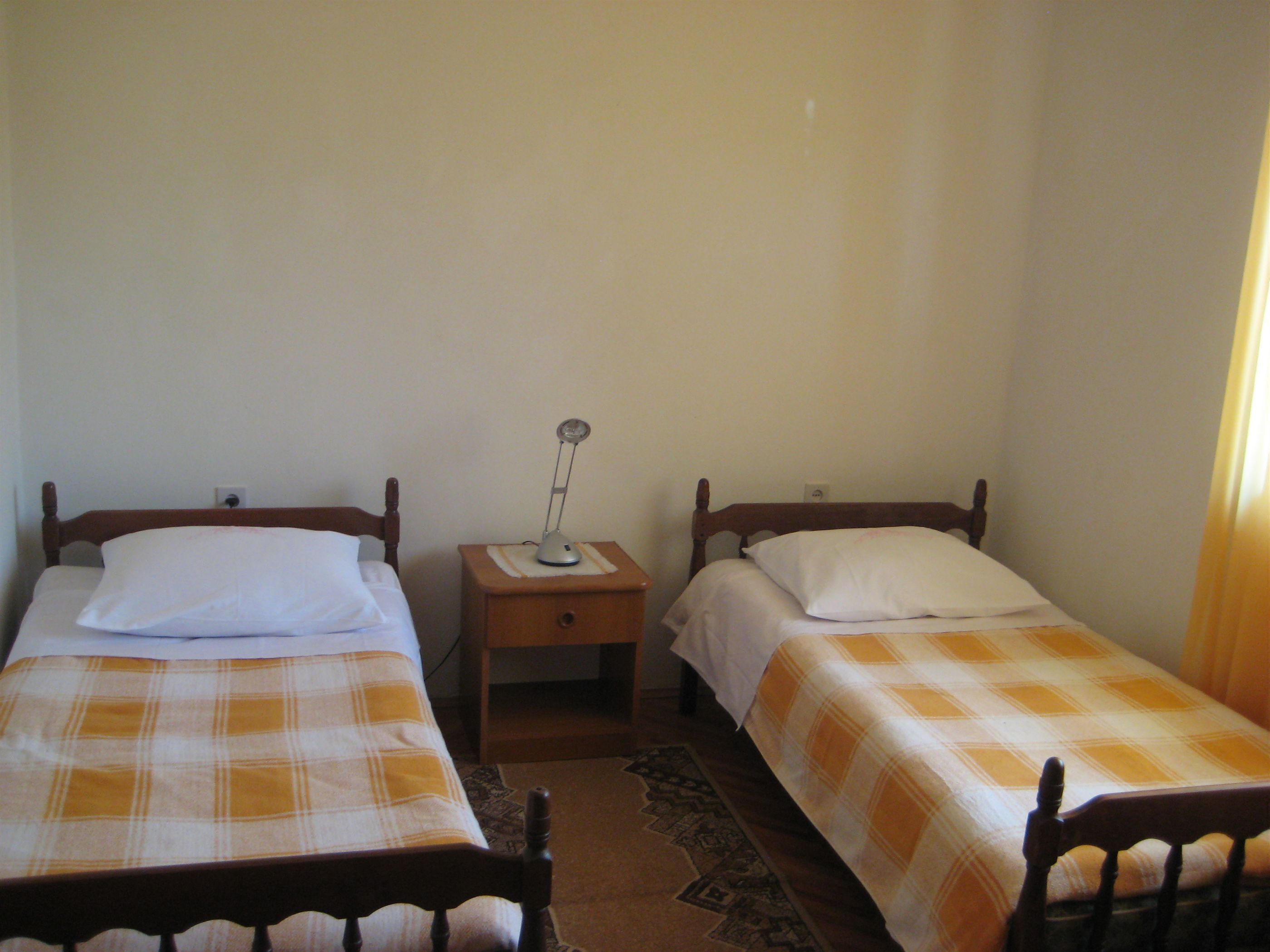 A room with two single beds, each with a wooden headboard and a plaid bedspread. There is a nightstand with a lamp between the beds.
