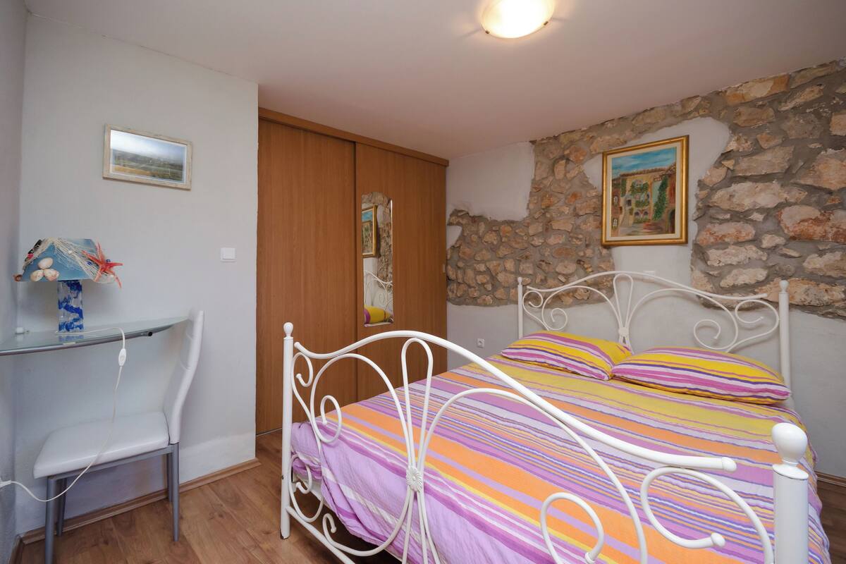 A bedroom with a bed, a desk, and a chair. There is a stone wall and a wooden door.