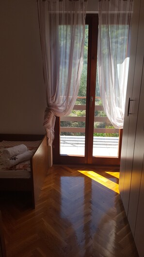 A room with a wooden floor, a bed with a floral bedspread, a sliding glass door with sheer curtains, and a view of greenery outside. - Apartment Ana (Kali)