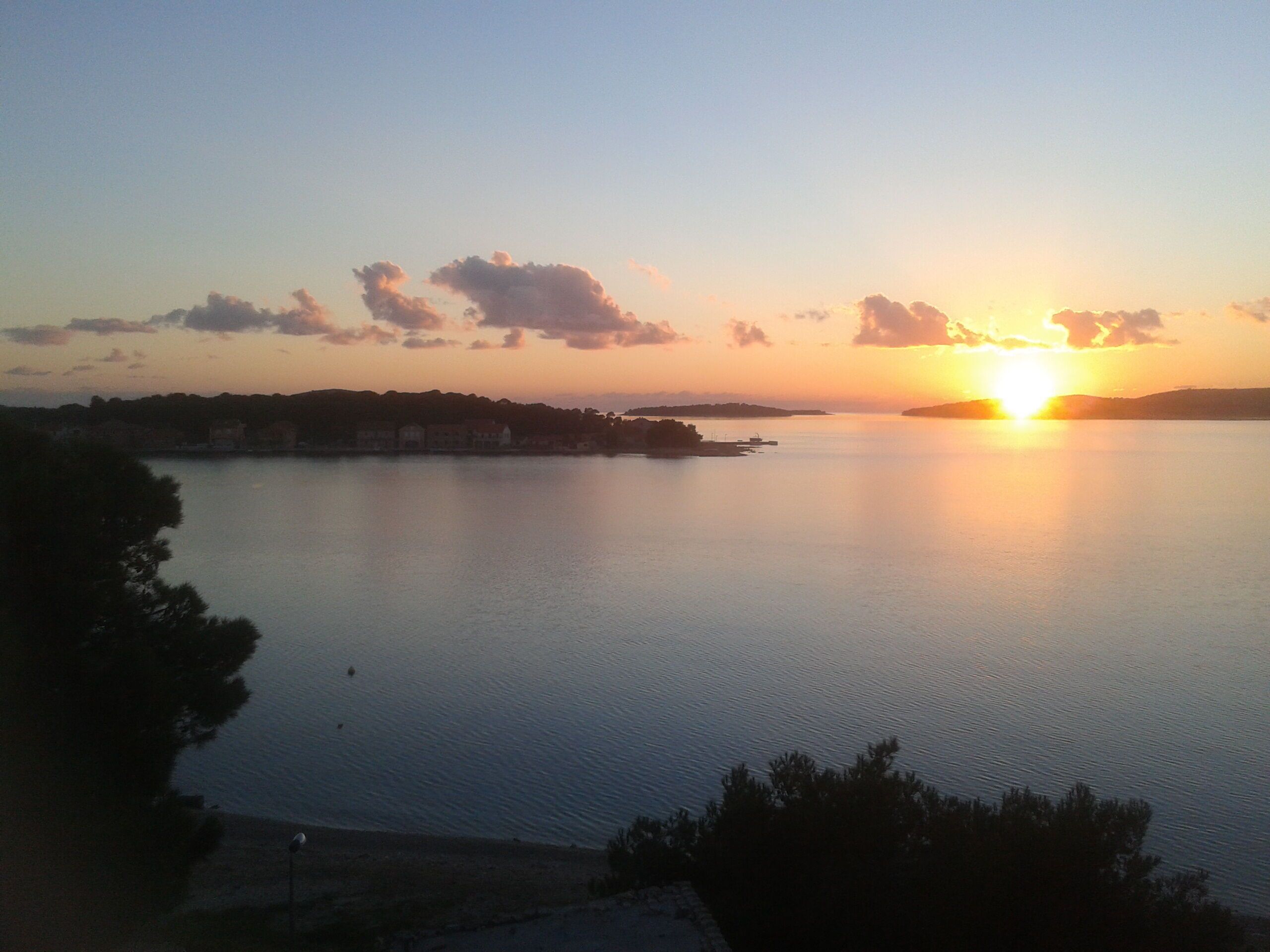 Un coucher de soleil sur une étendue d’eau calme, avec une côte et des bâtiments visibles au loin.