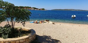 A pebble beach with a clear blue sea, a few boats, and a tree in the foreground. - Apartment Ivana (Sibenik)