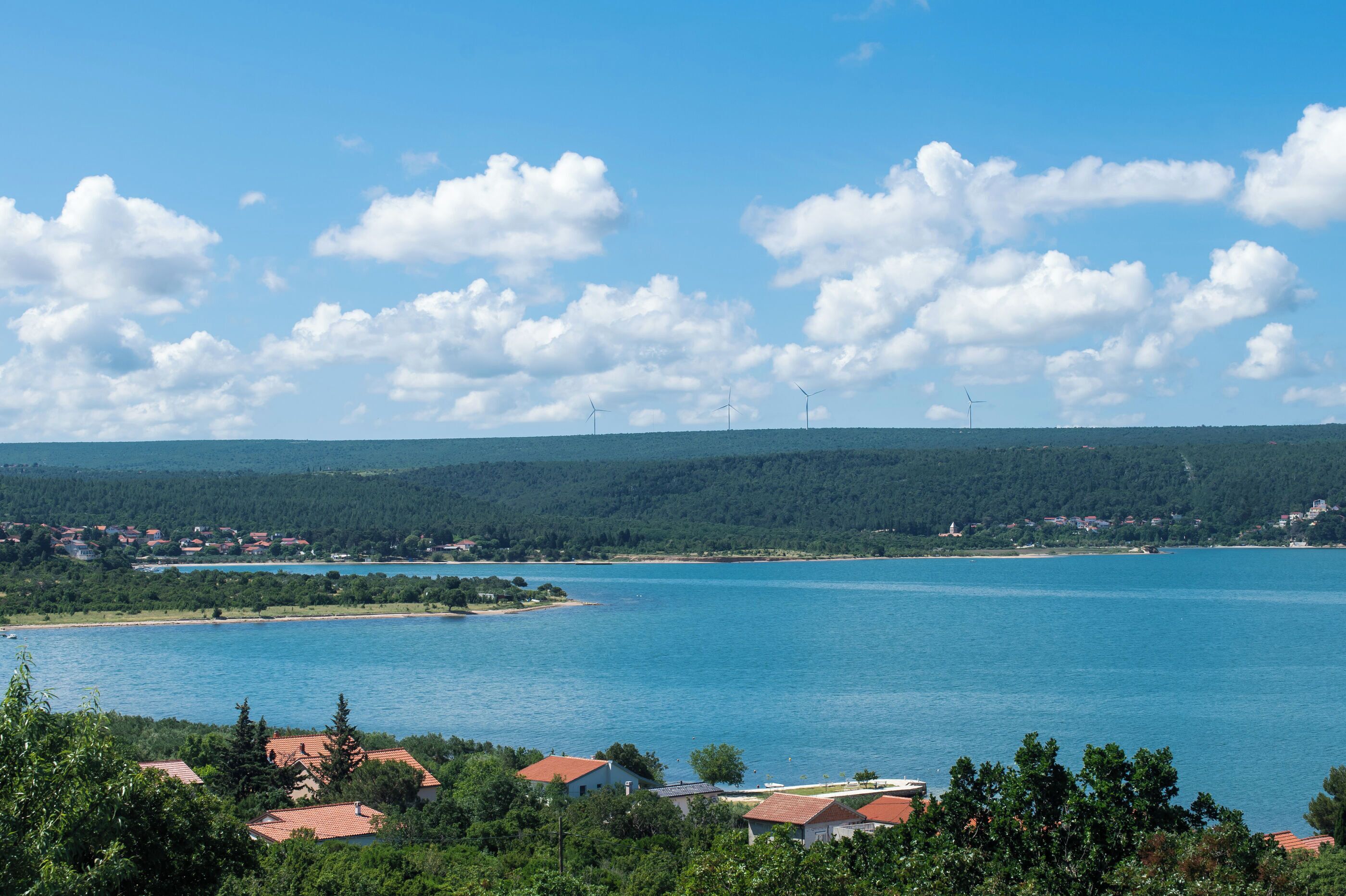 A coastal landscape with a clear sky, a calm sea, and a forested hill in the background.