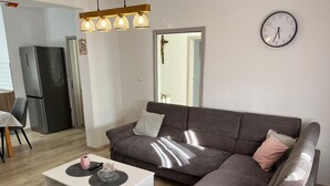A modern living room with a sectional sofa, a white coffee table, a wall clock, and a kitchen area in the background. - House Sea and River (Obrovac)