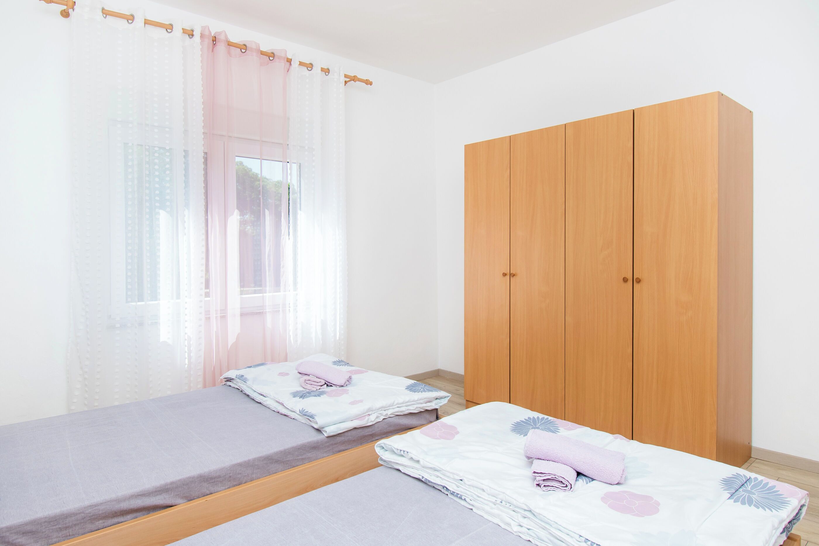 A bedroom with two single beds, a wooden wardrobe, and sheer curtains.