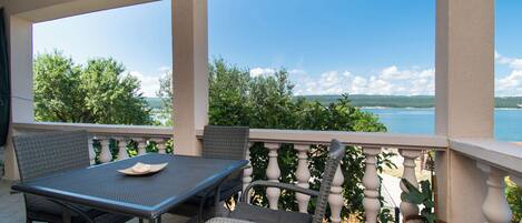 A balcony with a table and chairs, a potted plant, and a view of the sea and greenery.