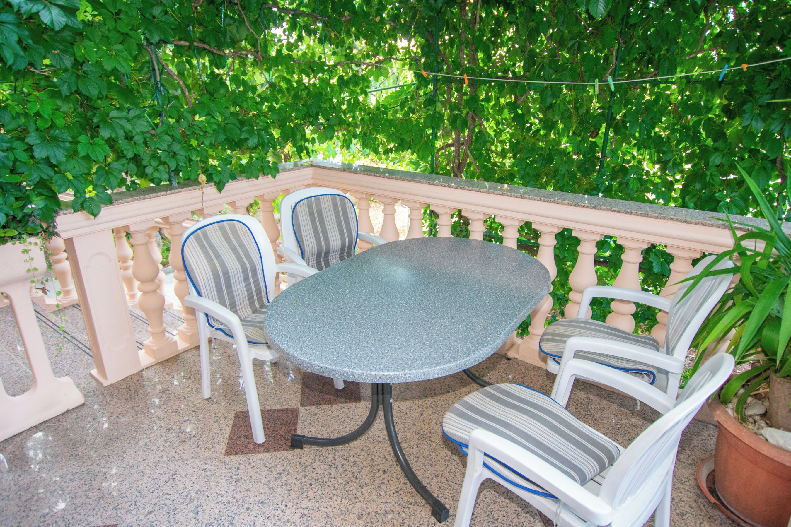 An outdoor patio with a round table and four striped chairs, surrounded by a white balustrade and lush green foliage.