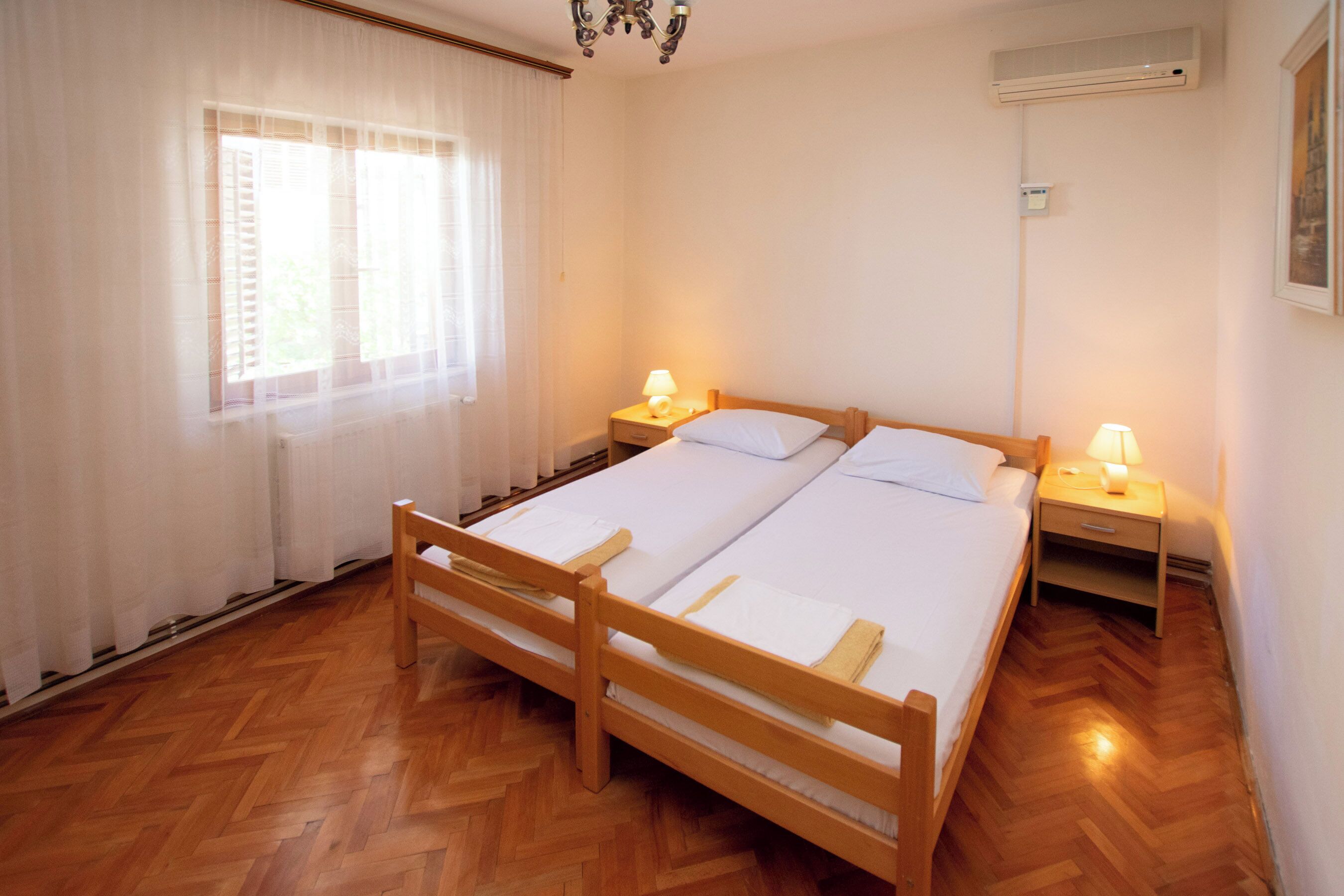 Una cama doble con cabeceros de madera, dos mesitas de noche con lámparas, una ventana con cortinas traslúcidas y un suelo de madera con estampado de espiga.