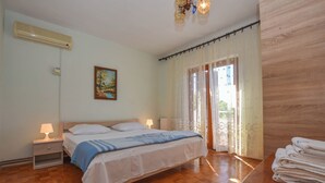 A bedroom with a wooden floor, a bed with white bedding, a nightstand with a lamp, a painting on the wall, and a window with curtains. - Apartments Immortelle (Biograd na Moru)