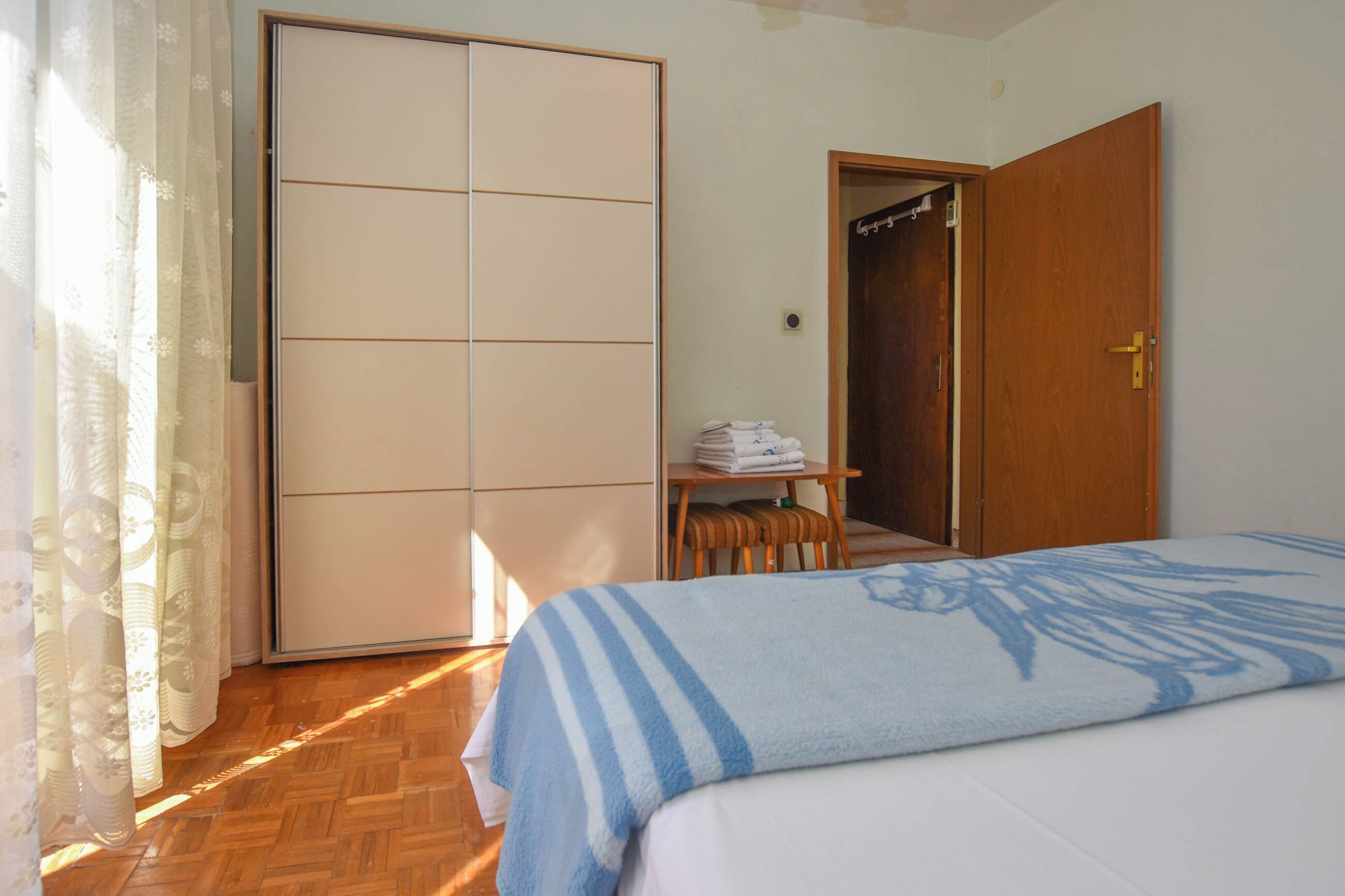 A bedroom with a bed, a wardrobe, a table with folded towels, and a door leading to another room.