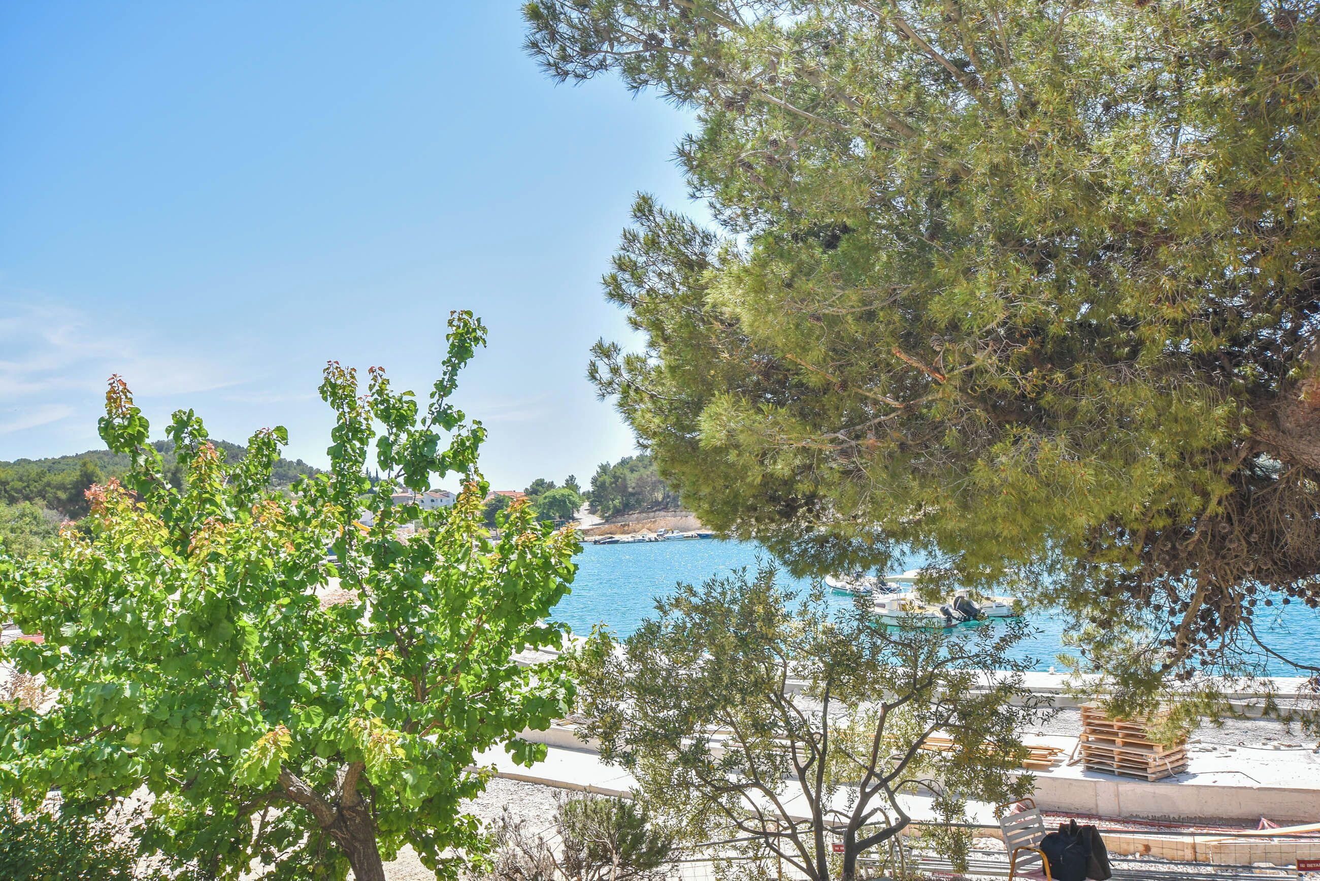 A coastal view with a clear blue sea, a sandy beach, and a few boats. There are trees in the foreground and a building with a balcony overlooking the sea.