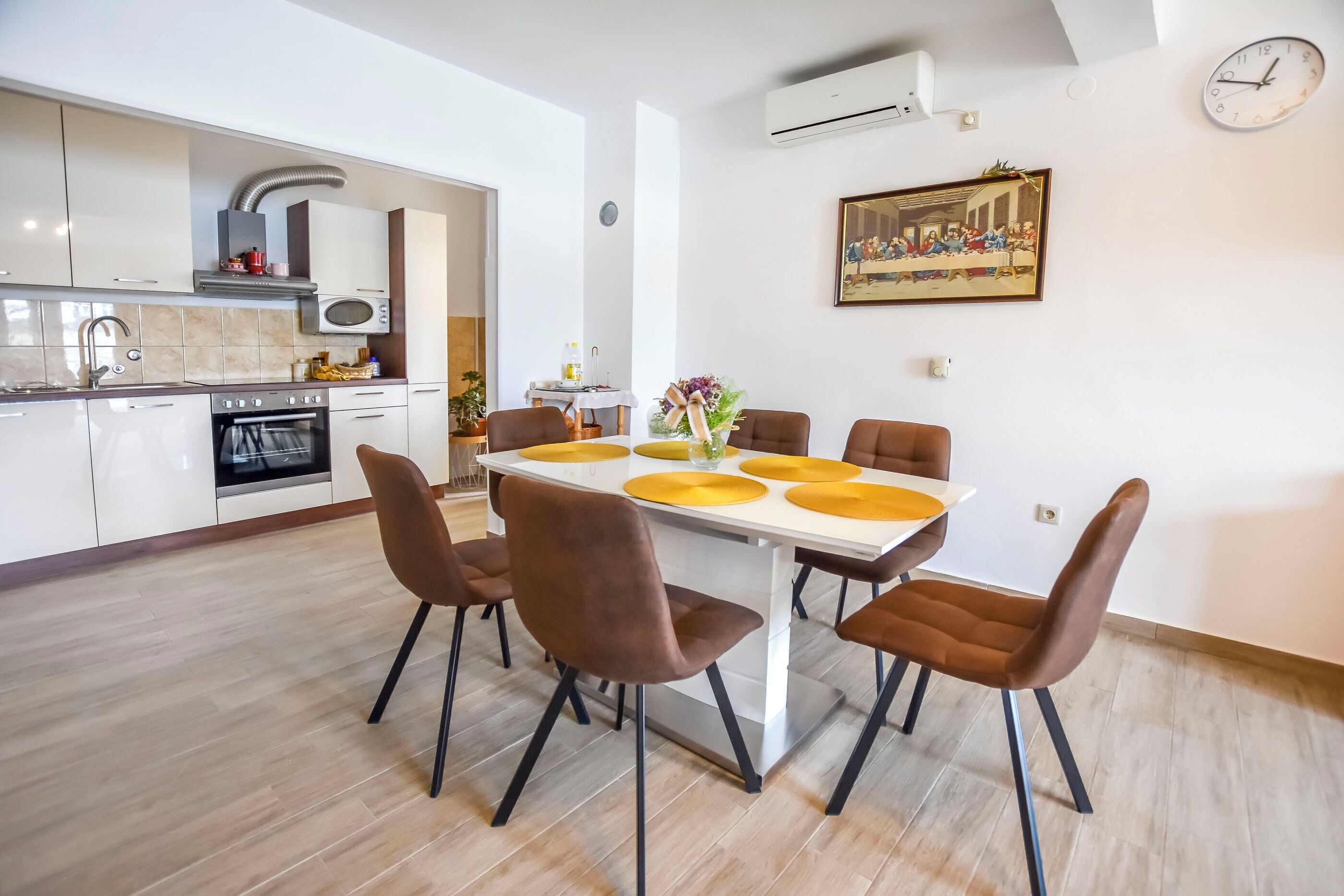 A modern kitchen with white cabinets, a microwave, and an oven. There is a dining table with brown chairs and yellow placemats. A clock and a framed picture are on the wall.