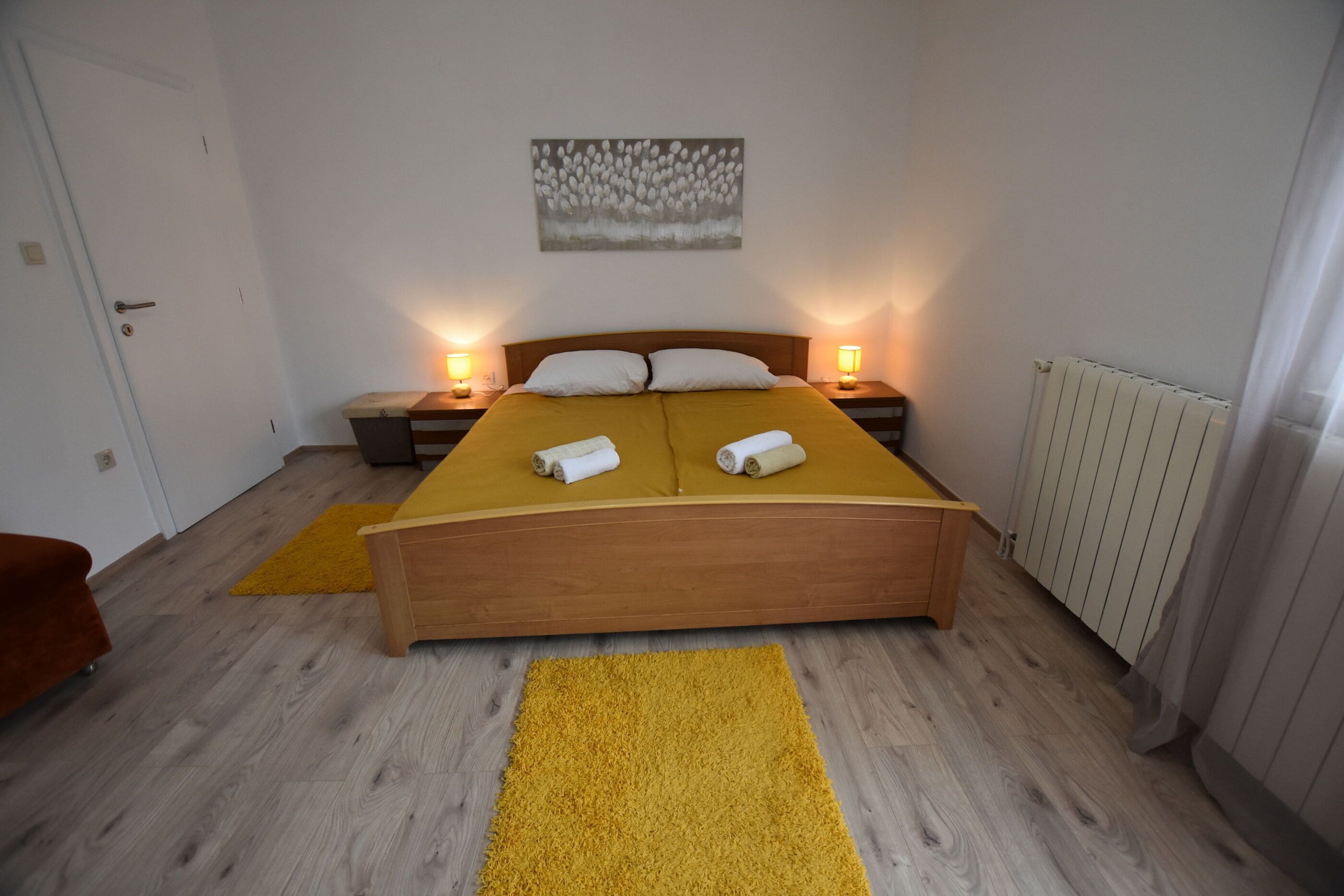 A bedroom with a wooden bed, two bedside tables with lamps, a radiator, and a yellow rug on the floor.