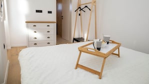 Una camera da letto con un letto, un comò in legno, un cavalletto di legno con vestiti e un vassoio con tazze e bicchieri.