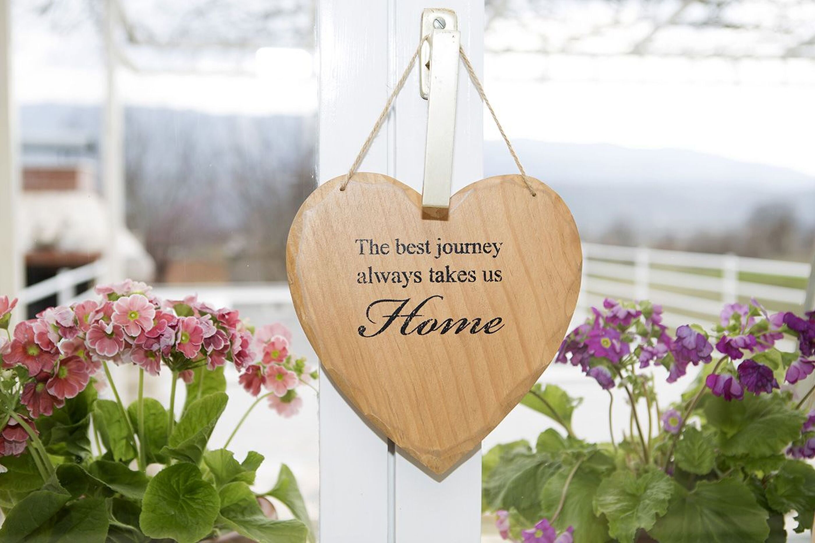 Ein hölzernes, herzförmiges Schild mit dem Aufdruck „The best journey always takes us Home“, an einem weißen Pfosten befestigt.