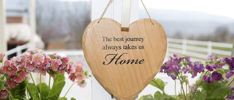 Ein hölzernes, herzförmiges Schild mit dem Aufdruck „The best journey always takes us Home“, an einem weißen Pfosten befestigt.