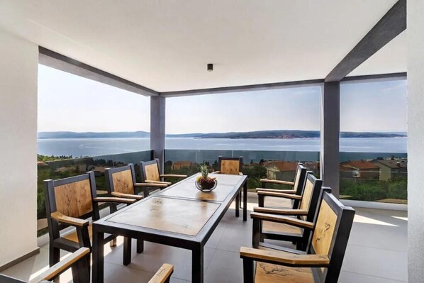 A dining area with a long table and chairs, large windows with a view of a body of water and landscape. - Villa Three Units (Crikvenica)
