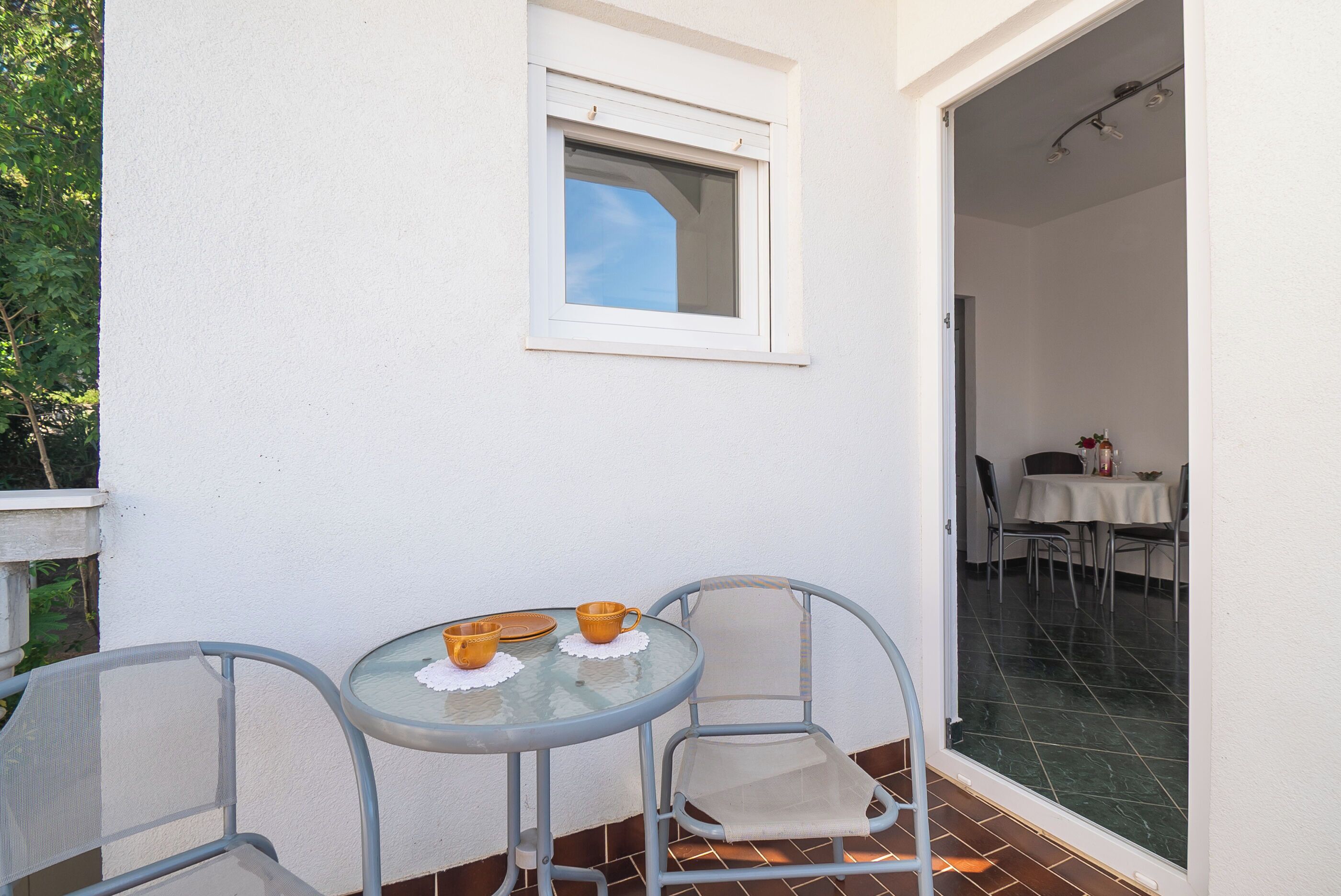 A small outdoor dining area with a table and two chairs, a window, and a view into a dining room.