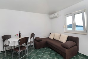A room with a brown sofa, a dining table with chairs, a bottle of wine, and a window with a view of the sea. - Apartments Branko (Tisno)