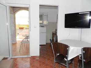 A dining area with a table and chairs, a television mounted on the wall, and a kitchen in the background. - Apartments Branko (Tisno)