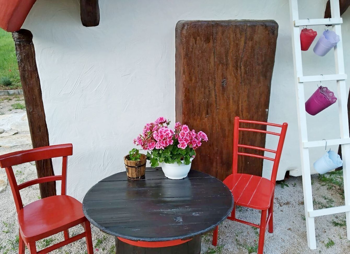 Ein runder Tisch mit zwei roten Stühlen, ein weißer Topf mit rosa Blumen und ein Holzbrett, das an einer weißen Wand lehnt.