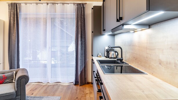Una cocina moderna con encimera de madera, estufa y hervidor. Hay una zona de comedor con sofá y ventana con cortinas.