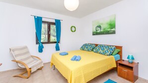 Un dormitorio con cama, una silla, una mesita de noche, una ventana con cortinas, un adorno de pared y una lámpara colgante.