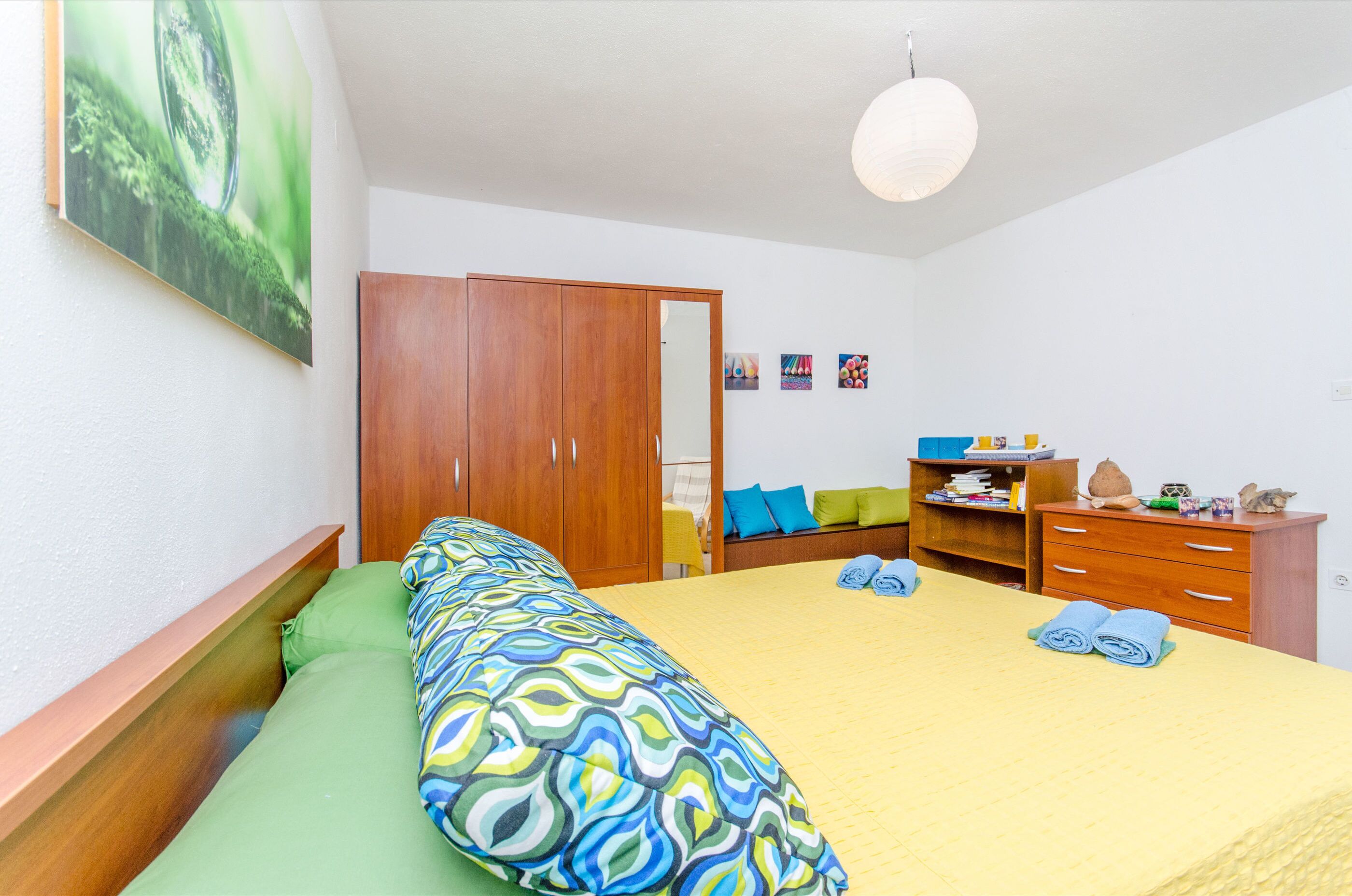 A bedroom with a bed, green and multicolored bedding, a wooden wardrobe, a desk with a chair, and a hanging light fixture.