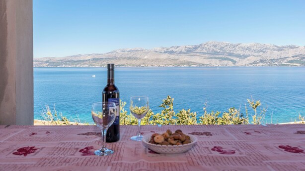 Un tavolo con una bottiglia di vino, un bicchiere e una ciotola di snack, con vista su un mare tranquillo e montagne in lontananza.