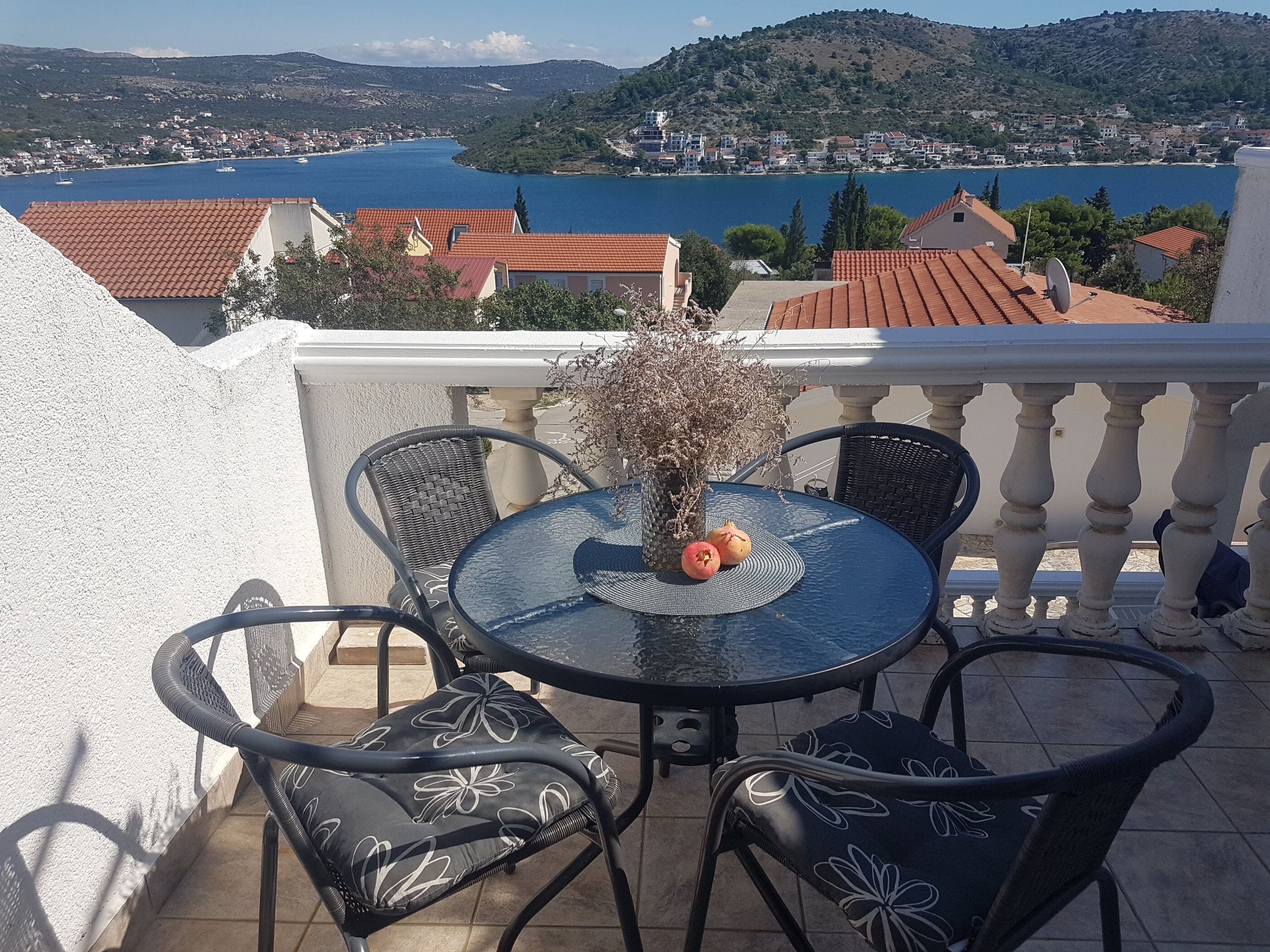 Un balcon avec une table ronde et des chaises, un pot de fleurs et une vue sur une ville côtière et la mer.