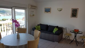 A room with a dining table, chairs, a sofa, a small table with a basket, and a view of the sea through a large window. - Apartments Adriatic (Rogoznica)