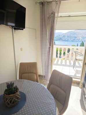 A balcony with a view of a body of water and hills, a round table with a polka dot tablecloth, two chairs, and a mounted TV. - Apartments Adriatic (Rogoznica)