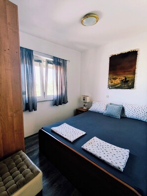 A bedroom with a large bed, a nightstand, a lamp, a window with curtains, and a painting on the wall. - Apartments Adriatic (Rogoznica)