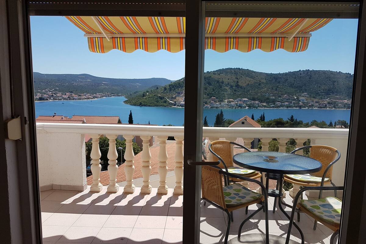 A balcony with a view of a lake and hills, featuring a table and chairs under striped awnings.