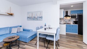 Una sala de estar moderna con un sofá azul, una mesa de comedor con cuatro sillas, una mesita de centro pequeña, un control remoto, una botella de vino y una copa. En la pared hay un cuadro con veleros y un modelo de barco en un estante. Se ve la cocina al fondo, con armarios azules y un microondas.