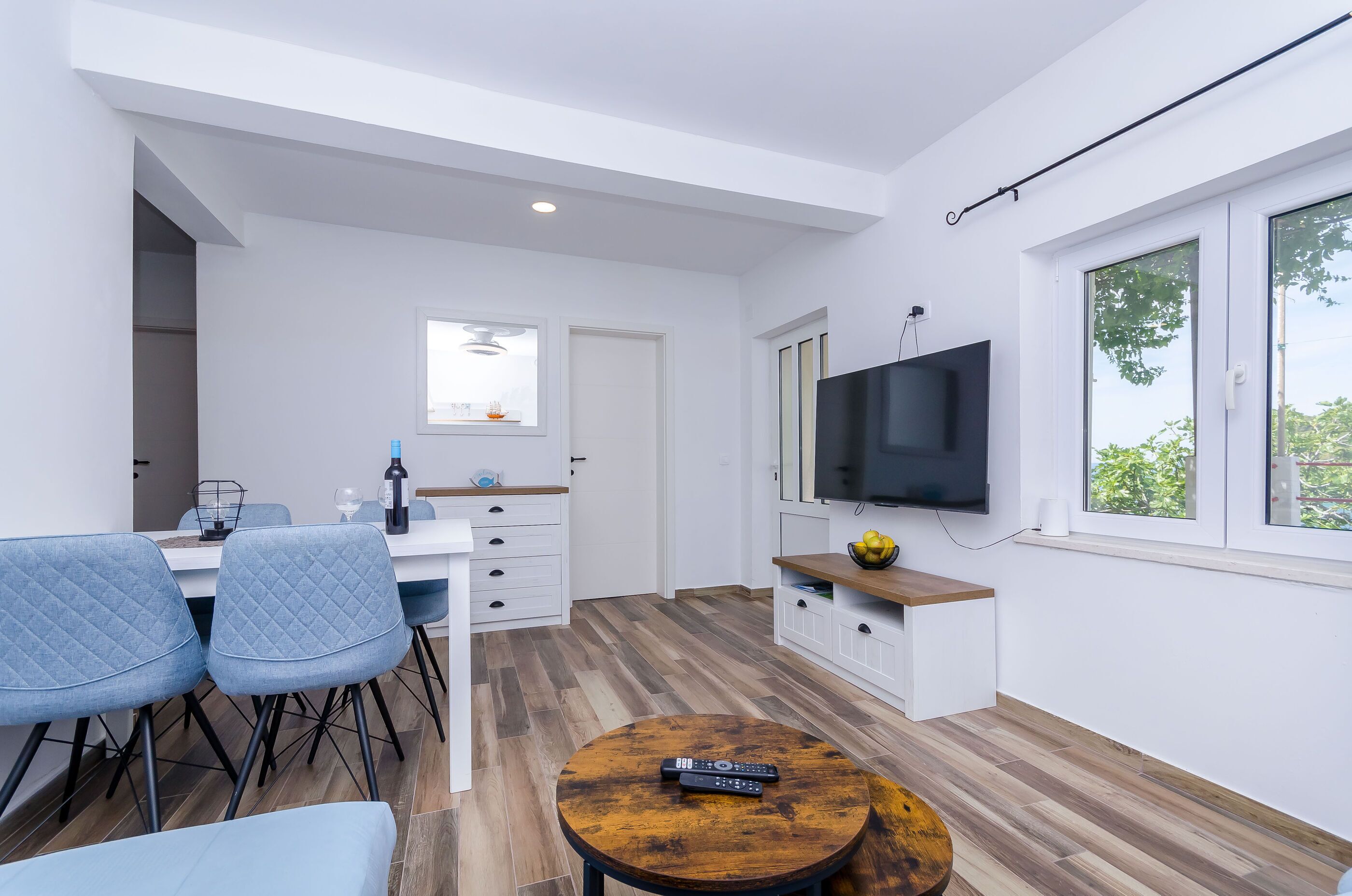 A modern kitchen with white cabinets, a dining area with blue chairs, a wooden coffee table, and a TV mounted on the wall.