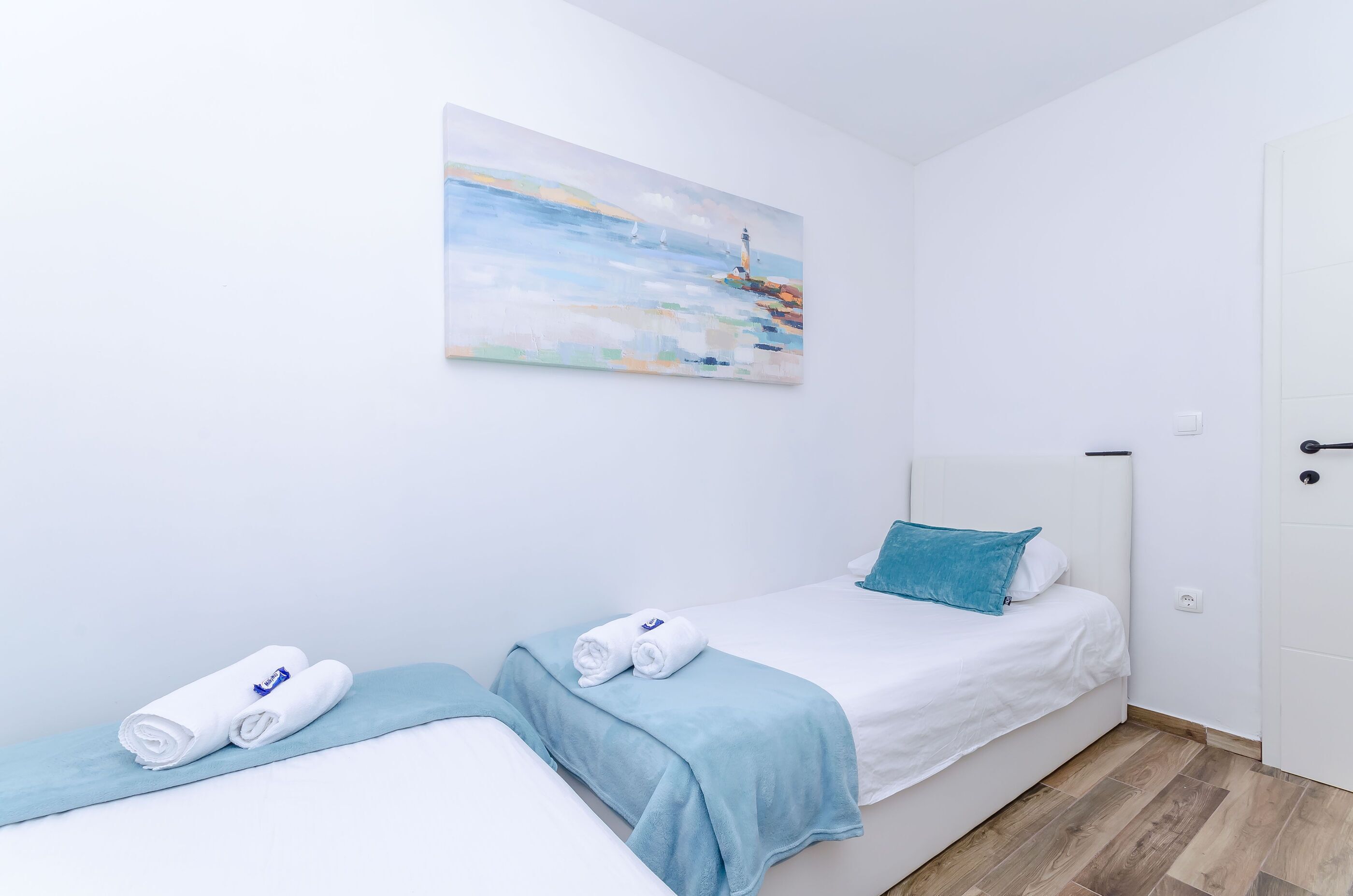 A hotel room with two beds, a painting of a lighthouse, and a blue towel on each bed.