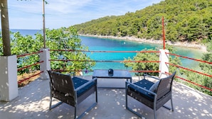 Un balcón con dos sillas de mimbre y una mesa, con vista al mar turquesa y una ladera cubierta de bosque.