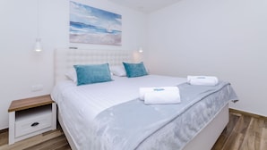 Una cama bien hecha con un cabecero blanco, dos almohadas color turquesa y una manta azul. En la pared sobre la cama hay un cuadro de un velero.