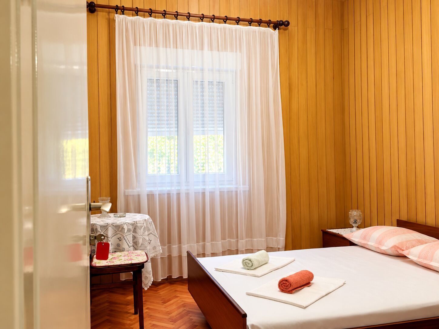 Una habitación de hotel con pared de madera, una cama con dos almohadas, una mesa con un vaso y una ventana con cortina.