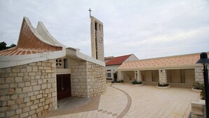 A courtyard with a church featuring a clock tower and a cross, surrounded by stone buildings with a tiled roof. - Apartments Soline (Podgora)