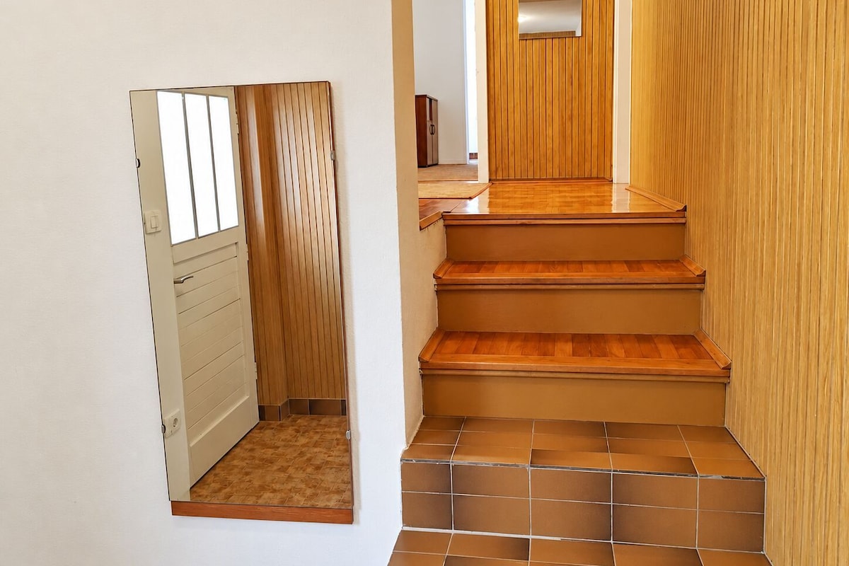 A narrow hallway with a wooden door and a small window, leading to a staircase with wooden steps and a tiled floor.