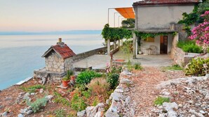 Ein Küstenhaus mit einem Steinsschornstein, einer Terrasse mit Gartenmöbeln und Blick auf das Meer.
