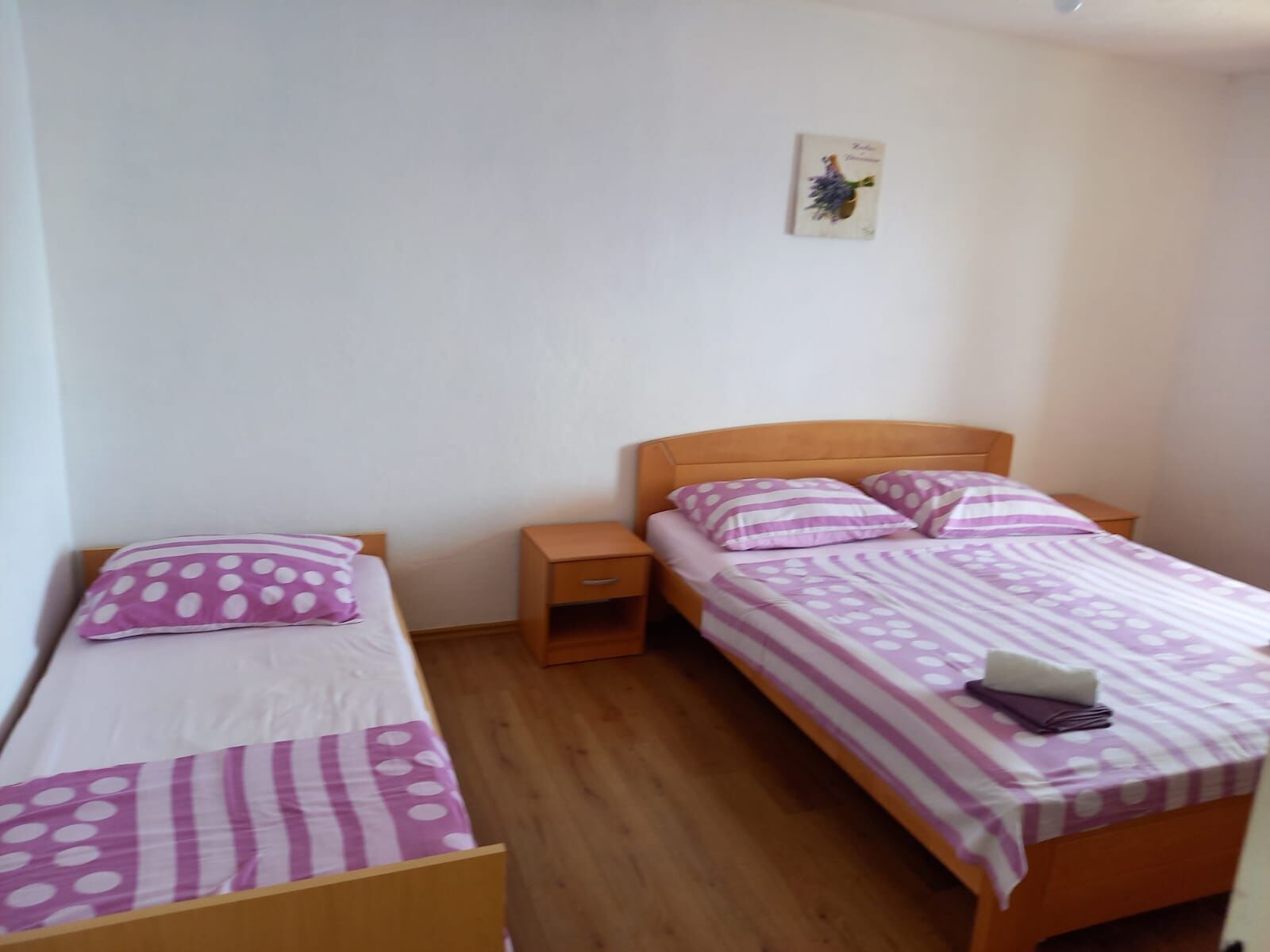 Quarto pequeno e limpo com duas camas de solteiro, cada uma com cabeceira de madeira e almofadas combinando. Há uma pequena mesa de cabeceira entre as camas e uma mesa pequena com um abajur. As paredes são brancas e lisas, e há um quadro na parede acima das camas.