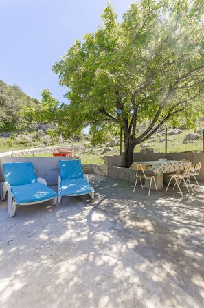 An outdoor seating area with blue lounge chairs and a table, surrounded by a stone wall and a tree. - House Sv. Lovre (Postira)