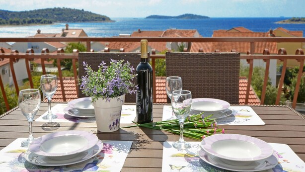 A table set for two with plates, glasses, a bottle of wine, and a potted lavender plant on a balcony overlooking a coastal view. - Apartments Petar (Rogoznica)