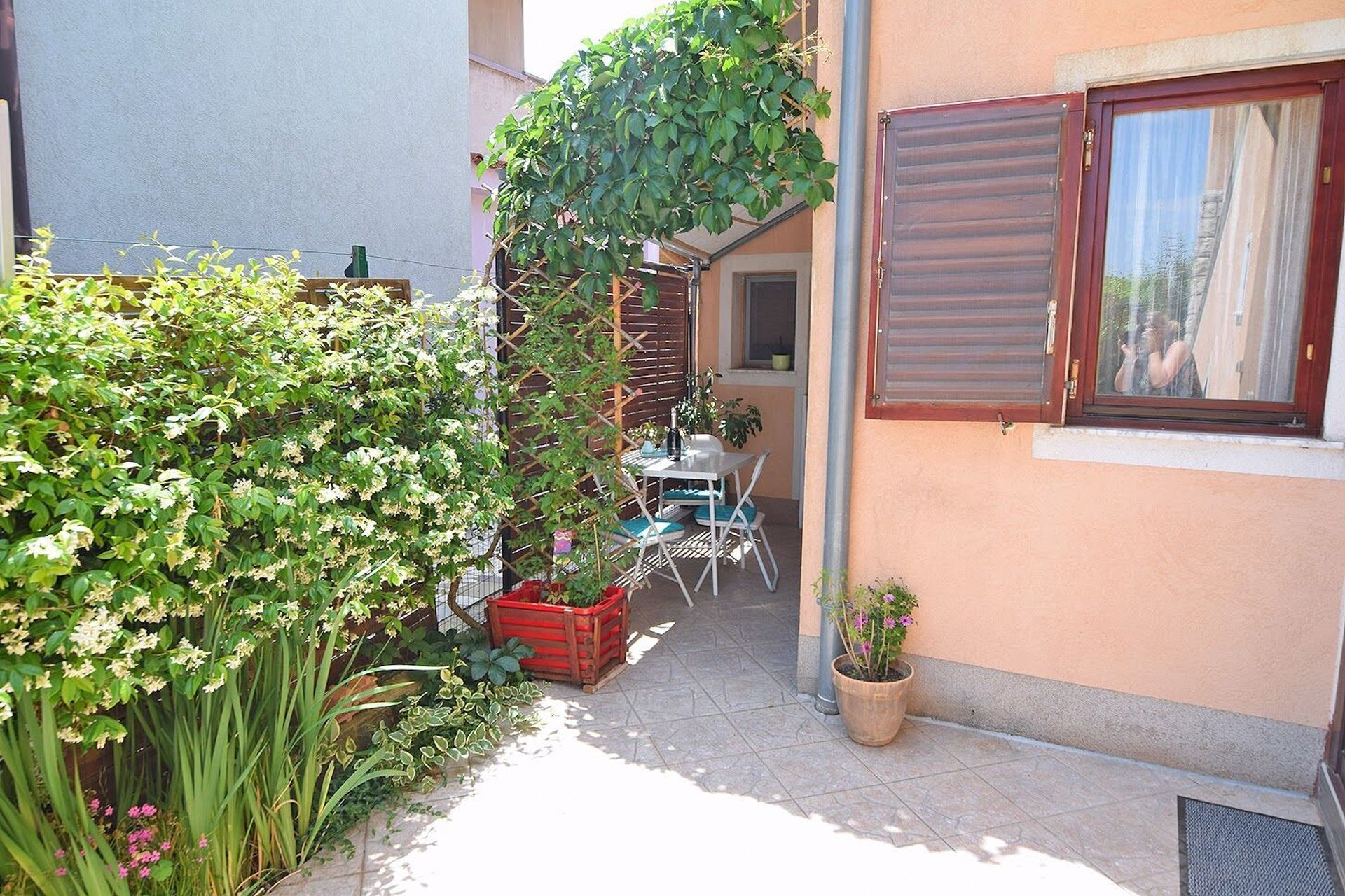 A narrow outdoor area with a table and chairs, a potted plant, and a window with shutters.